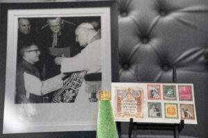 A golden ring on a stand in the foreground. A stamp book and photo of Bishop Malone and the pope in the background.