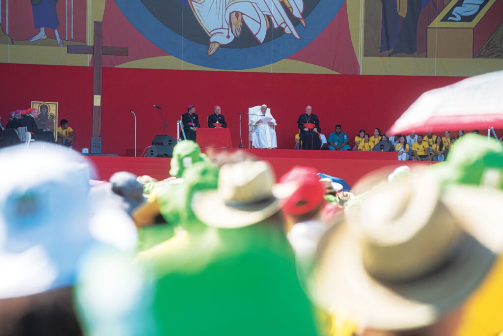 Pope Francis is seated on a stage amdist a massive crowd.